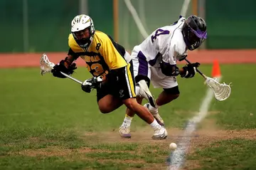 A Man wearing a yellow and a black sport jersey holding lacrosse sticks.